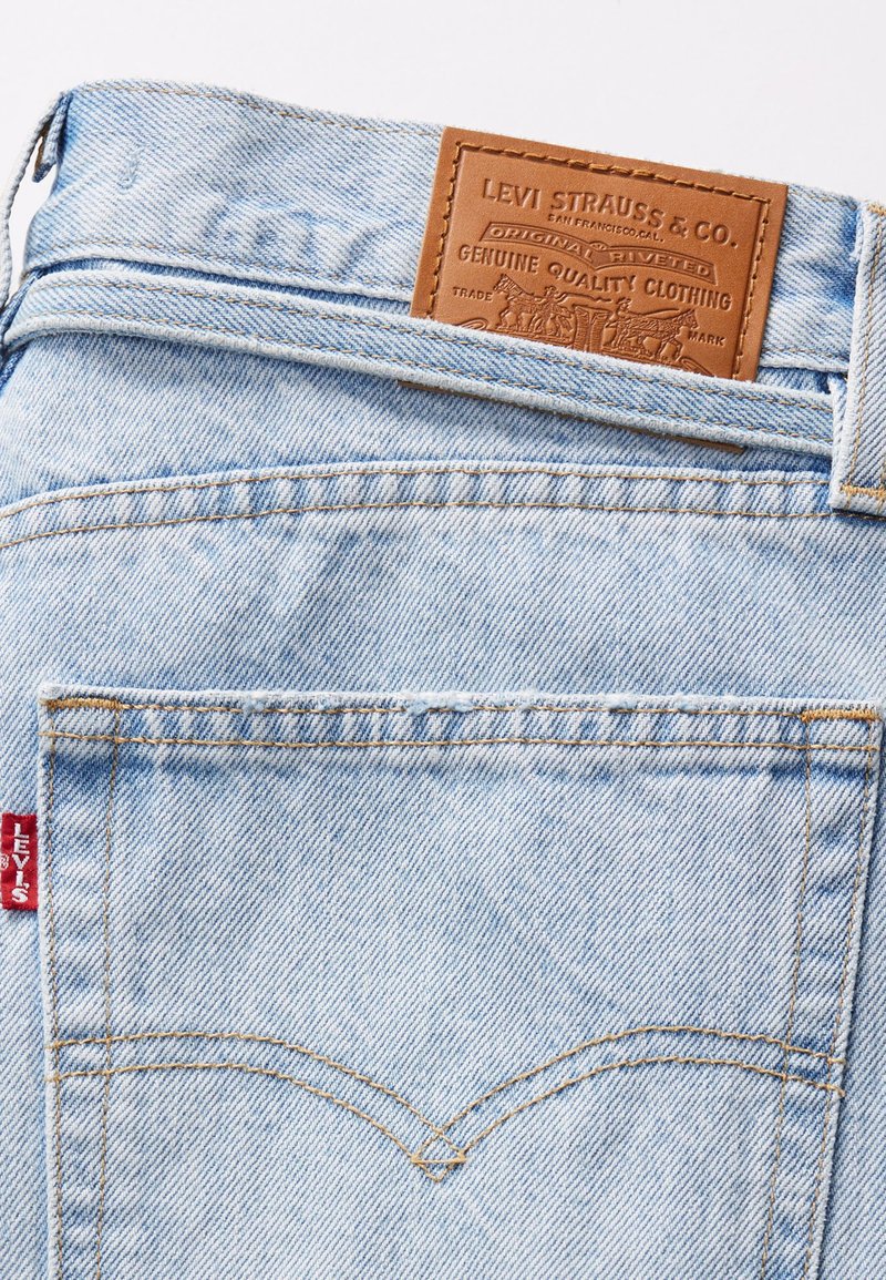 Un jean en denim bleu clair présente une étiquette en cuir portant la mention "Levi Strauss & Co." avec des détails de couture et une petite étiquette rouge sur le côté.