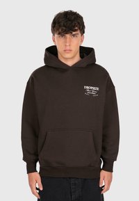Un jeune homme aux cheveux bouclés porte un sweat à capuche marron foncé avec le texte "DROPSIZE Street Game" sur la poitrine et un pantalon noir, se tenant devant un fond uni.