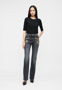 Vrouw staat, draagt een zwart shirt met korte mouwen, donker hoge taille jeans met gouden slangenborduursel op de heupen en zwarte puntige schoenen tegen een witte achtergrond.