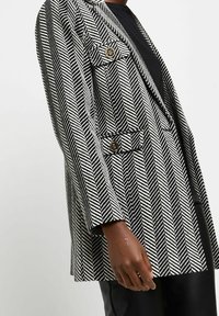 Sako s herringbone vzorem v černobílé barvě, zlaté knoflíky a klopový límec, se uvolněným střihem a mírně oversized rukávy.