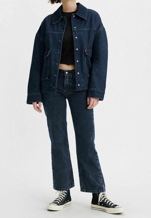 Persoon die een donkerblauw spijkerjack en spijkerbroek draagt, een zwart cropped top en zwarte hoge sneakers met witte veters, staand op een witte achtergrond.