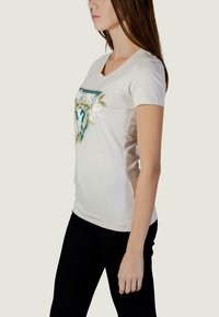 T-shirt crème à manches courtes, col en V, avec un motif floral et un point d'interrogation central. Associé à un pantalon noir ajusté.