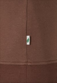 Brun genserstoff med ribbet kant og et lite hvitt merke med et grønt bladdesign. Jevn tekstur med synlig søm.