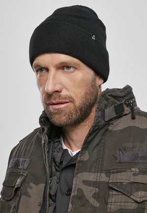 Zwart gebreid beanie op een kortgeknipt hoofd, gecombineerd met een camouflage-jack dat patches en een ritsdetail heeft.