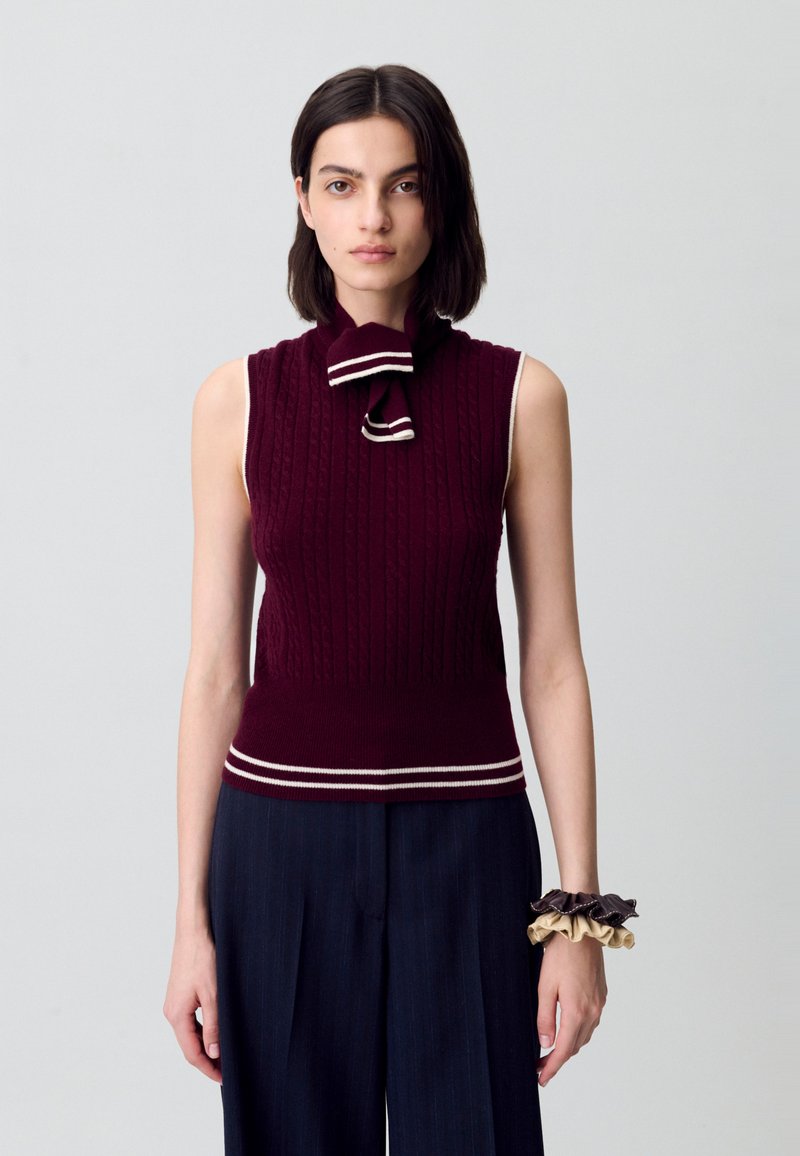 Top de punto sin mangas en burdeos con patrón de cable y franja blanca en el dobladillo, con un cuello de lazo. Combinado con pantalones de pierna ancha en azul marino.