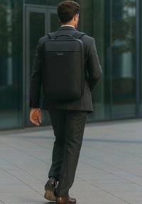 Mochila negra con diseño minimalista, hecha de material suave, que cuenta con un asa superior y correas ajustables, llevada por un hombre con traje.