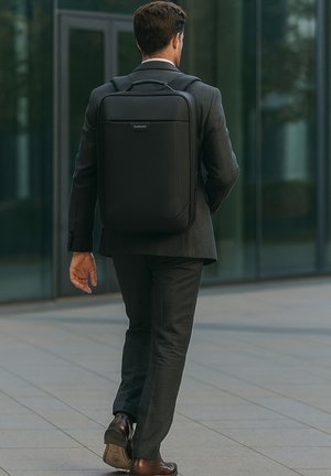 Un sac à dos noir au design minimaliste, fabriqué en matériau lisse, avec une poignée supérieure et des bretelles réglables, porté par un homme en costume.