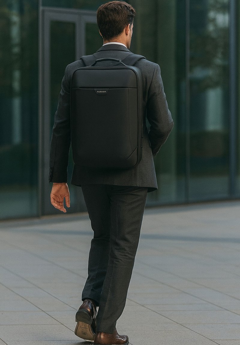 Mochila negra con diseño minimalista, hecha de material suave, que cuenta con un asa superior y correas ajustables, llevada por un hombre con traje.