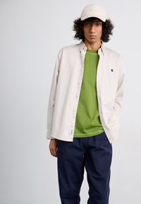 Cămiză deschis bej cu nasturi peste un tricou verde, asortată cu pantaloni închiși la culoare, navy și o șapcă bej, având un logo pe cămașă și pe șapcă.