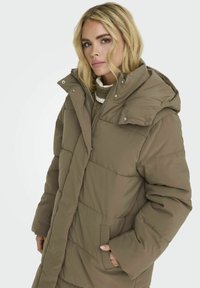 Manteau long rembourré en olive avec un col montant, fermeture éclair et boutons-pression ; il présente des coutures diagonales et des poches latérales.