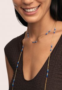 Collana in oro con perline blu e azzurro chiaro, caratterizzata da un design delicato e schemi di perline alternati, indossata sopra una maglia scura con texture.