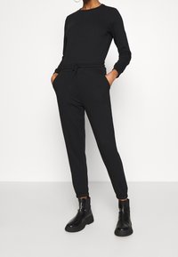 Mono negro de manga larga, con cintura elástica y cordón. Presenta piernas cónicas y bolsillos laterales. Se combina con botas negras de tobillo.