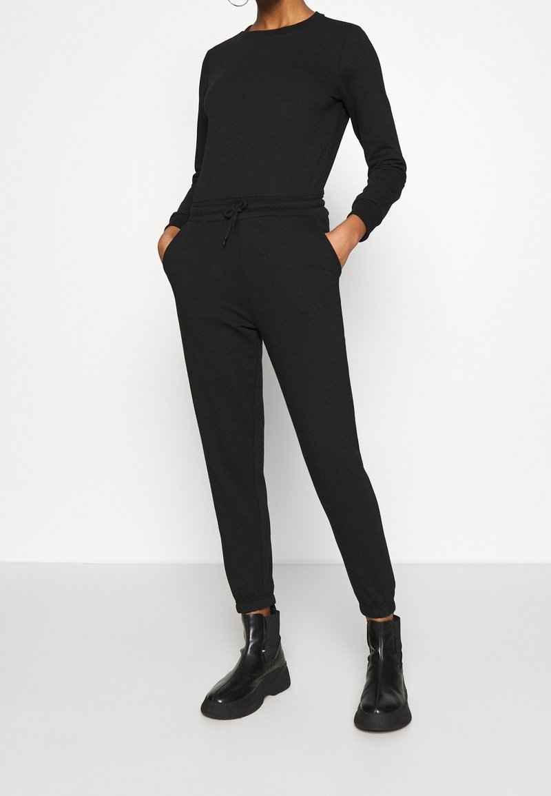 Mono negro de manga larga, con cintura elástica y cordón. Presenta piernas cónicas y bolsillos laterales. Se combina con botas negras de tobillo.