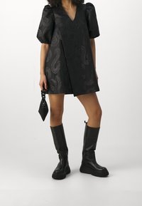 Schwarzes kurzes Kleid mit Puffärmeln, V-Ausschnitt, strukturiertem Muster, kombiniert mit kniehohen schwarzen Stiefeln und einer kleinen, strukturierten Tasche.