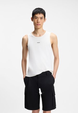 Hombre joven vistiendo una camiseta sin mangas blanca Hugo y pantalones cortos cargo negros, de pie con las manos en los bolsillos frente a un fondo liso.