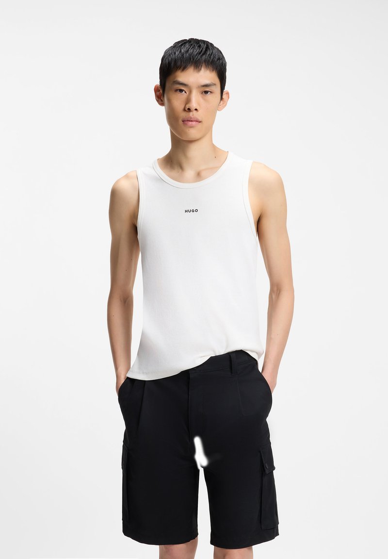Jeune homme portant un débardeur blanc Hugo sans manches et un short cargo noir, debout les mains dans les poches devant un fond uni.