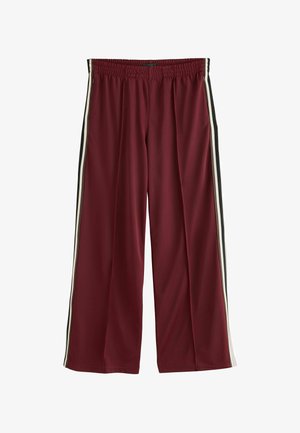 Kvinde iført brun jakke, hvid tanktop, bordeaux vidde bukser, beige sko, holder brun håndtaske mod en ensfarvet beige baggrund.