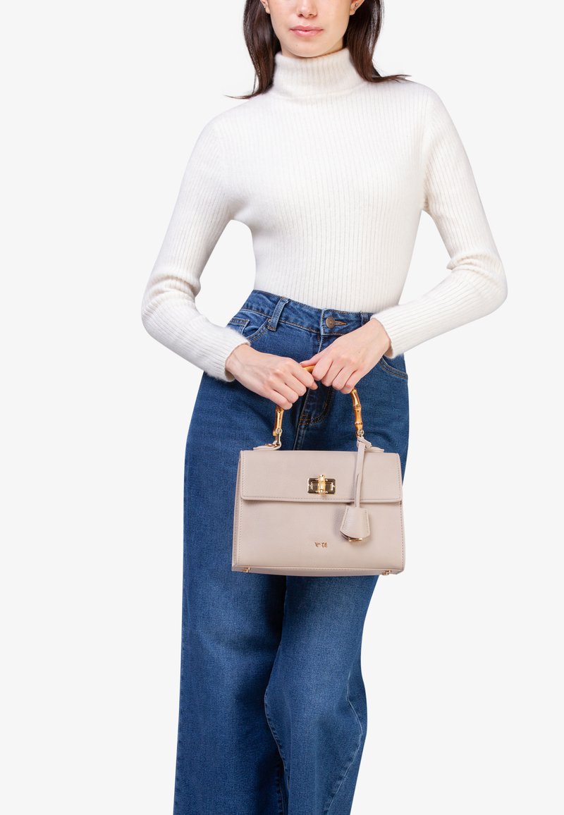 Bolso de cuero beige con un diseño estructurado, herrajes dorados y una etiqueta desmontable. El modelo lo sostiene con un asa de madera natural.