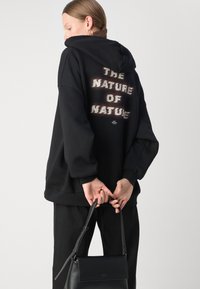Svart oversized hettegenser med teksten "NATURAS NATUR" på baksiden. Kombinert med svarte bukser og en liten svart lærveske.