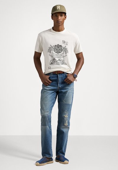 Balts kokvilnas T-krekls ar drukātu grafiku, zili denima džinsi ar nolietotiem pleķiem un zili espadrilles. Olīvu krāsas cepure ar logo akcentu.