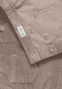 Jasnofioletowa tkanina kordurowa z teksturową powierzchnią, z zapięciami na guziki i przeszytymi kieszeniami. Widoczne są akcenty w kolorze srebrnym.