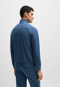 Giacca sportiva blu con colletto alto, dotata di maniche lunghe, una consistenza liscia e piping a contrasto lungo le spalle.