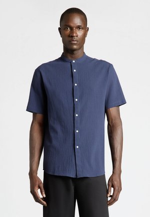 Man wearing a short-sleeve, navy blue textured shirt with white buttons standing against a plain light background.