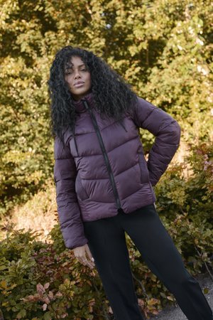 Opblaasjack in diep paars met een hoge kraag, ritssluiting en gestructureerde zijpanelen. Gepaard met zwarte broeken. Groene loofachtergrond.