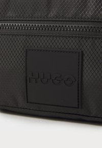 Nahaufnahme eines schwarzen, strukturierten Stoffes mit einem Reißverschluss und einem schwarzen rechteckigen Patch, auf dem das Wort "HUGO" geprägt ist.