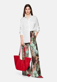 Weißes Hemd mit Knöpfen, florale weit geschnittene Hose in Brauntönen, Grün und Rot, und eine rote strukturierte Tote-Bag. Rote spitze Schuhe sind sichtbar.