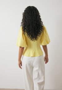 Gelbes Oberteil mit Puffärmeln und taillierter Form, das eine glatte Textur und ein strukturiertes Design aufweist, kombiniert mit weißen weiten Hosen.