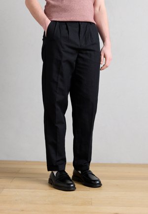 Personne portant un pantalon noir plissé, des mocassins en cuir noir, des chaussettes claires et une chemise rose à manches courtes rentrée, debout sur un sol en bois.