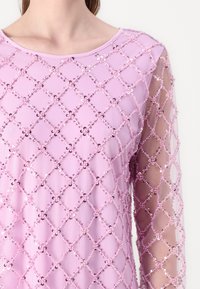 Roze top met een met pailletten versierde diamantpatroon, doorschijnende lange mouwen en een ronde halslijn. De stof is glad met een gestructureerde afwerking.