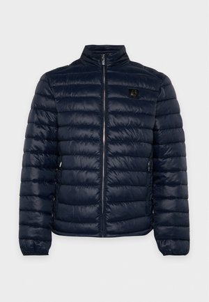 Un homme porte une veste matelassée bleu marine sur une chemise et un pantalon noirs, avec des bottes noires, devant un fond uni et clair.
