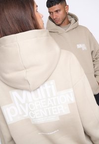 Sudadera beige de tejido suave con un gran texto en blanco que dice "Centro de Creación Múltiple". Diseñada con una capucha amplia y un bolsillo tipo canguro.