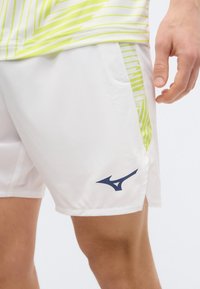 Άνδρας που φοράει λευκά αθλητικά σορτς με πλαϊνές λωρίδες σε σχέδιο πράσινο λάιμ και λογότυπο Mizuno σε ναυτικό μπλε χρώμα στο αριστερό πόδι.