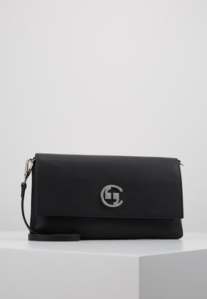 Bolso de mano de cuero negro con correa de muñeca desmontable y emblema de logo circular plateado en el frente, exhibido sobre un pedestal blanco contra un fondo gris.