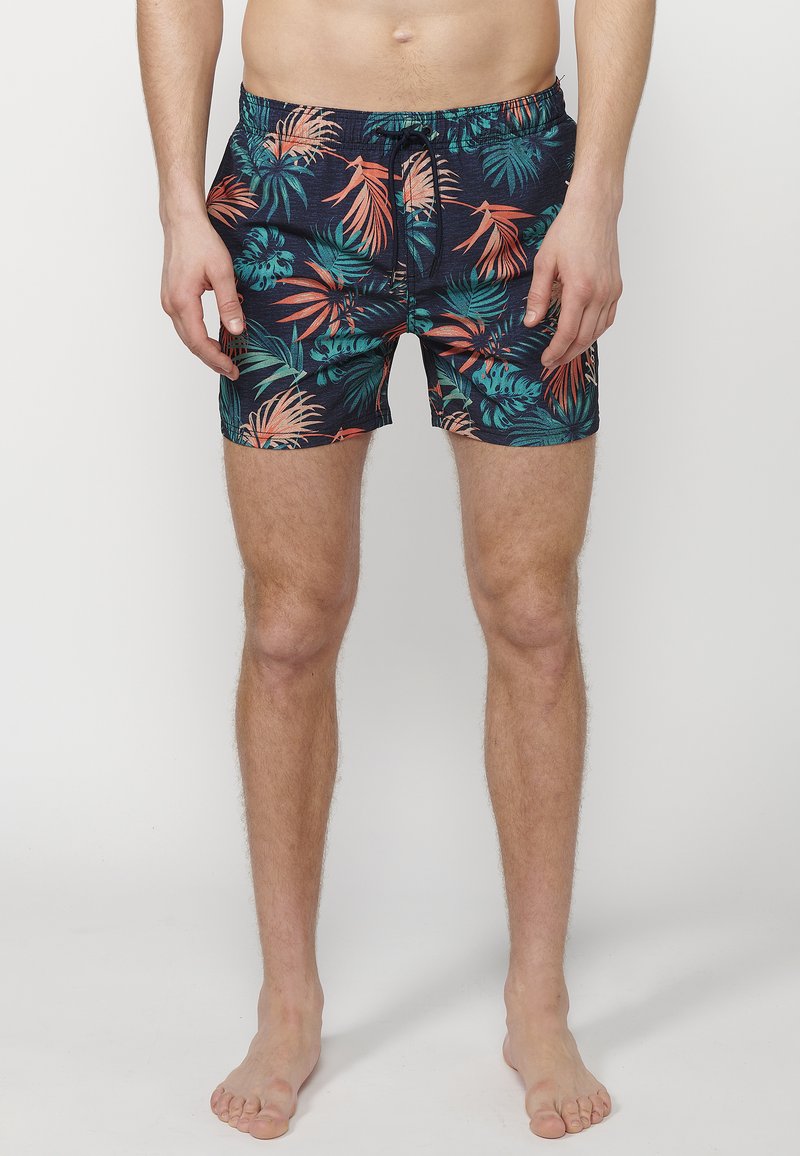 Costume de baie scurt, de culoare bleumarin, cu un model de frunze tropicale în verde și corai. Talie elastică cu șnur, lungime scurtă și material moale.