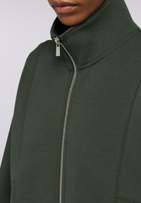 Zelená mikina s vysokým límcem, obsahující dlouhý stříbrný zip a jemné detaily stehu. Vyrobena z měkkého, texturovaného materiálu.