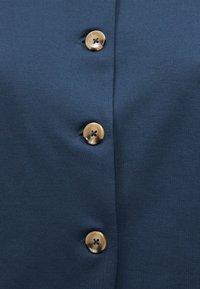 Ύφασμα σε ναυτικό μπλε με τρία ανοιχτό καφέ κουμπιά με τέσσερις τρύπες, ραμμένα σε κάθετη γραμμή, το καθένα στερεωμένο μέσω κουμπότρυπας.