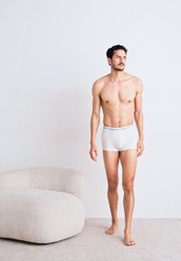 Boxer-briefs en coton blanc avec une ceinture bleu clair, portés par un modèle masculin la peau nue debout sur un tapis doux et de couleur claire.