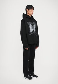 Melna pārspīlēta kapuce ar tauriņa grafiku un tekstu "VERSACE JEANS COUTURE", apvienota ar melnām biksēm un melniem sporta apaviem ar baltajiem akcentiem.