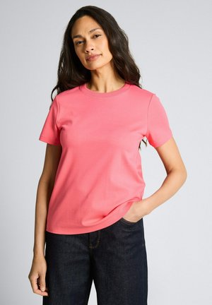 Frau mit langen dunklen Haaren, die ein korallenrotes kurzärmeliges T-Shirt und dunkle Jeans trägt, mit einer Hand in der Tasche vor einem schlichten Hintergrund stehend.