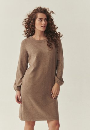 Kobieta z kręconymi włosami, ubrana w brązową sukienkę z długim rękawem sięgającą kolan, patrząca w bok na jednolitym tle.