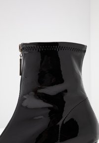 Bota de tobillo de charol negro con cremallera lateral y detalle de costura en zigzag en el borde superior, contra un fondo blanco liso.