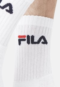 Chaussette blanche à la cheville avec une texture côtelée. Présente un logo Fila en noir, souligné de rouge au niveau de la cheville.