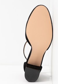 Sandale noire avec semelle beige, talon bloc et bride à la cheville vue de dessous sur fond blanc.