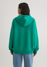 Green oversized hoodie with a drawstring hood, ribbed hem, and cuffs. The fabric appears soft and textured, paired with light blue jeans.