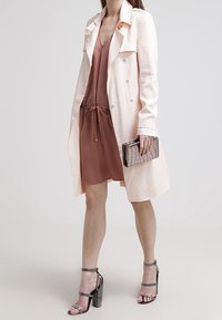 Jasnoróżowy płaszcz trencz na brązowej sukience, trzymająca srebrną kopertówkę o fakturowanej powierzchni. Sandały na wysokim obcasie o metalicznym wykończeniu. Gładka tkanina i dopasowany krój.