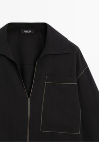 Chemise noire avec coutures blanches contrastantes, col pointu et grande poche avant sur le côté gauche, étiquetée Massimo Dutti.